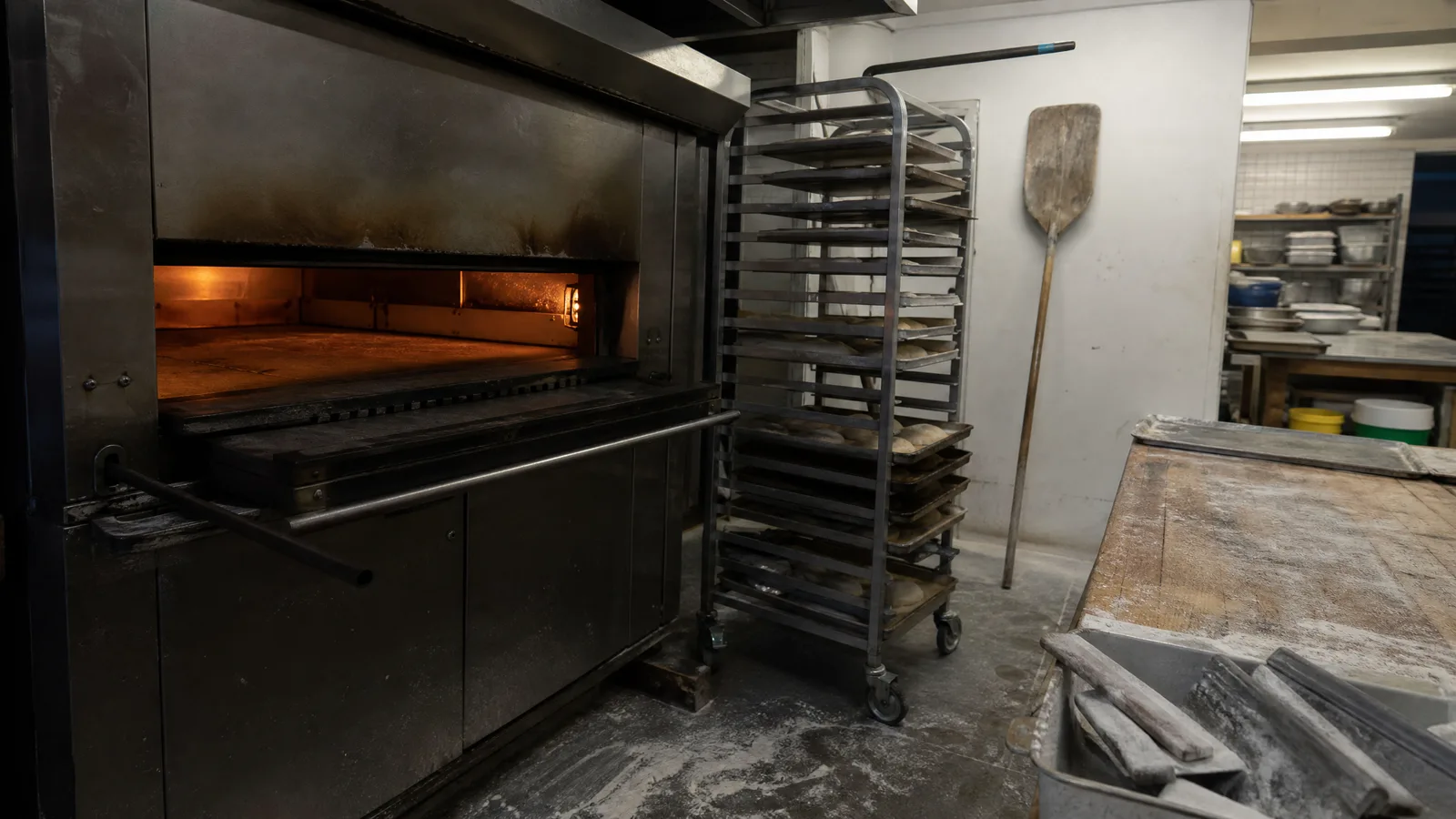 Open bakery deck oven beside a rolling rack of sheet trays in a working back-of-house bakery