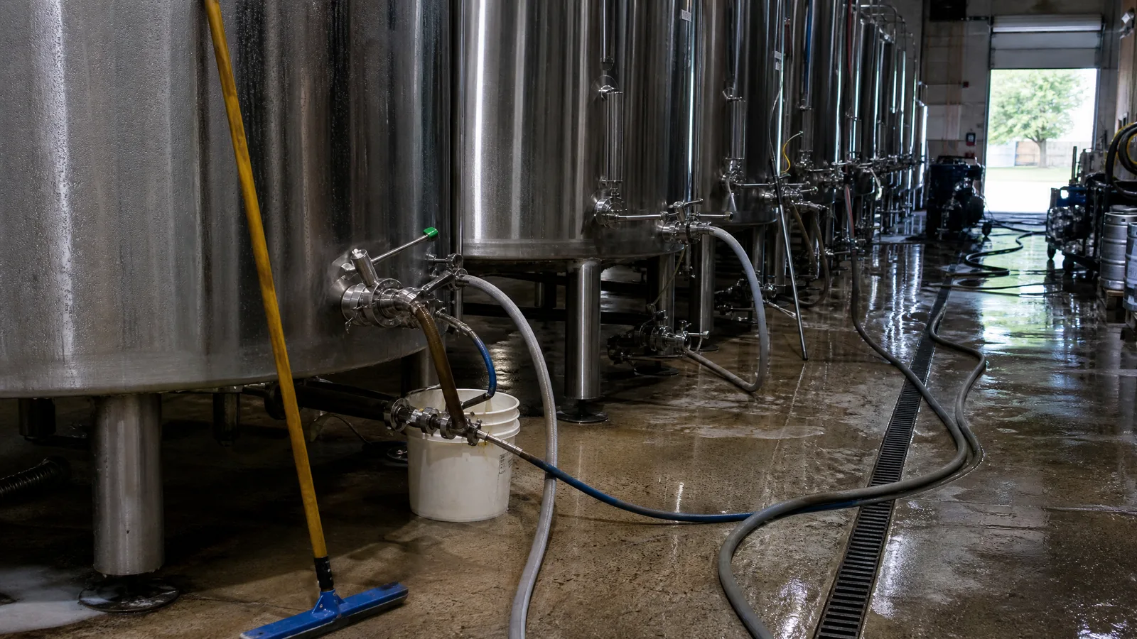 Row of stainless brite tanks in a brewery with hoses run across a wet sloped floor to a drain