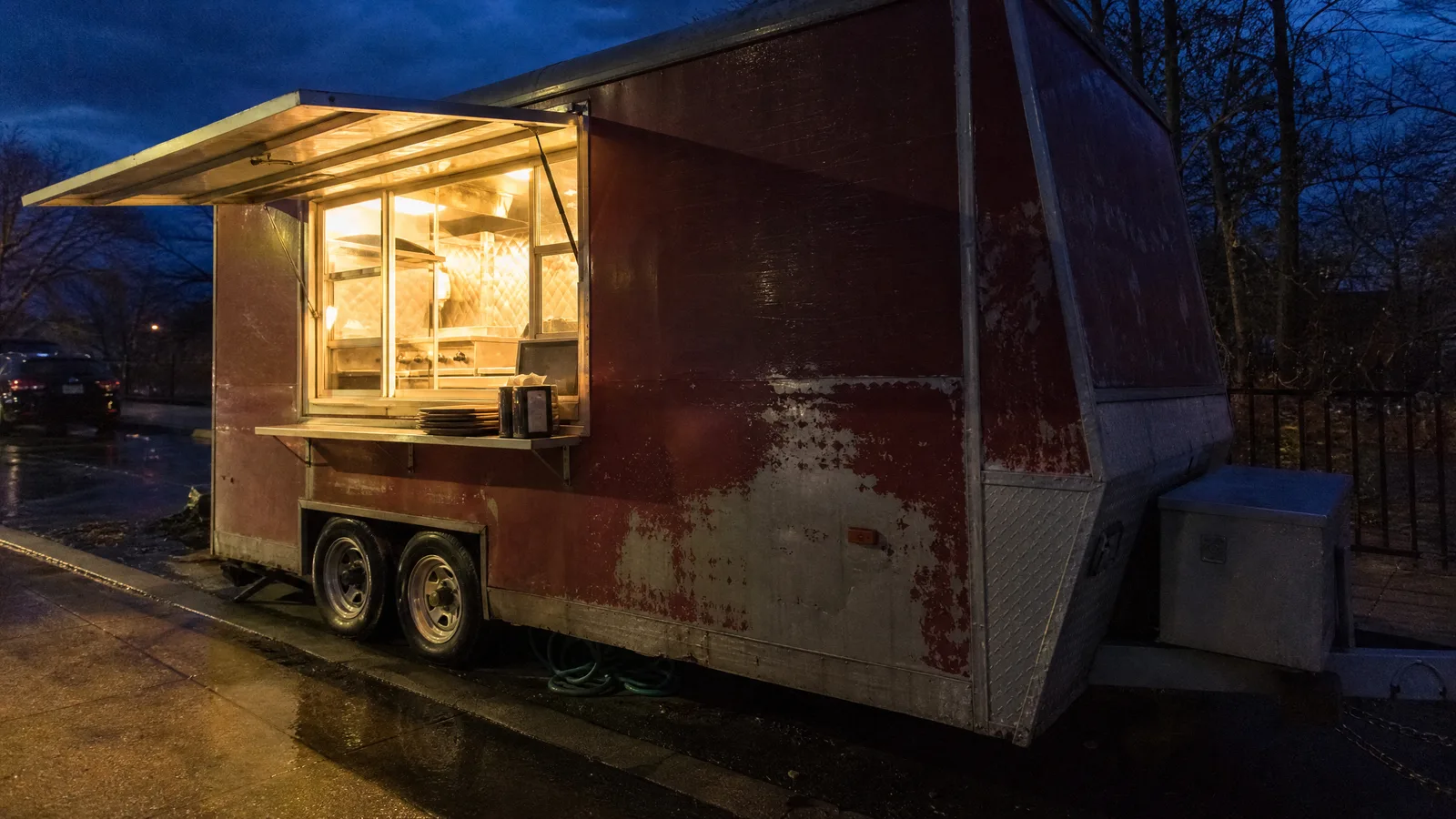 Food truck with its service window open and lit from inside, awning down, parked on a street at dusk